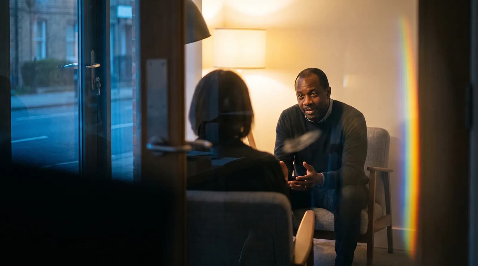 Through a consulting-room door, a practitioner leans forward to ask a question, the other person seen only from behind.