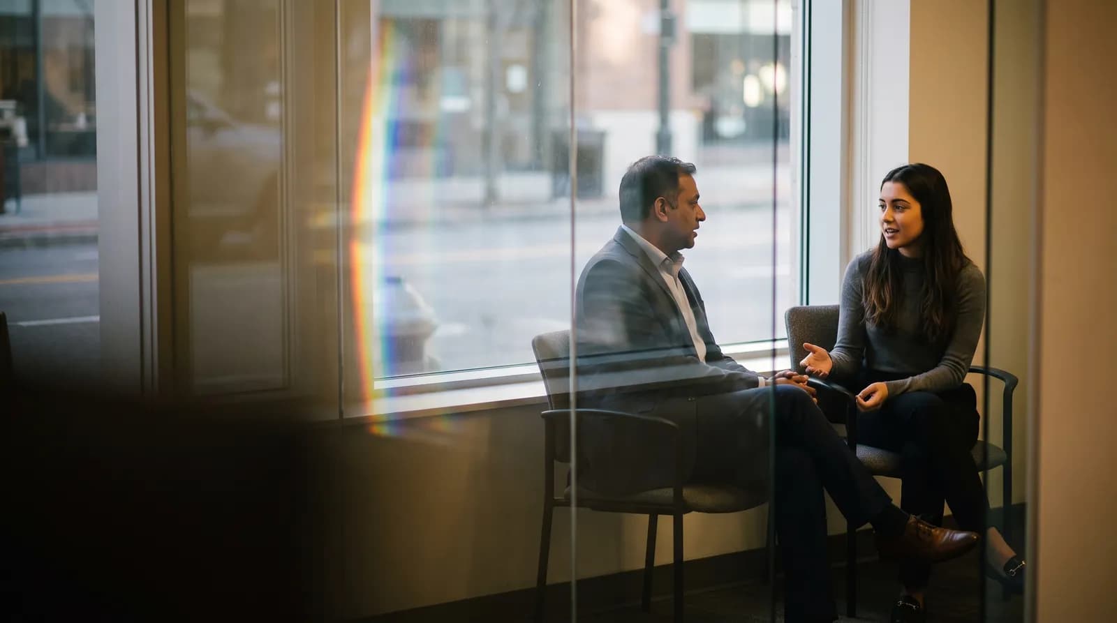 A manager and a colleague deep in a one-on-one by a window, seen through the office's glass door.